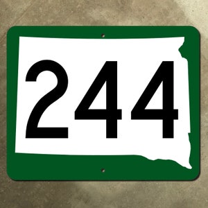 May include: Green and white sign with the number 244 on it. The sign is shaped like the state of South Dakota.