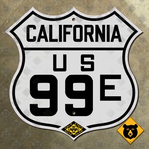 Pode incluir: Um sinal de metal branco com texto preto e borda. O sinal diz "California US 99E". Um pequeno adesivo amarelo em forma de diamante com um urso preto está no canto inferior direito. O sinal tem um furo na parte superior para pendurar.