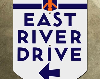 Könnte beinhalten: Ein weißes Schild mit blauem Text und einem blauen Pfeil, der nach links zeigt. Das Schild lautet "EAST RIVER DRIVE". Die Oberseite des Schildes hat ein blaues und orangefarbenes Streifenmuster.