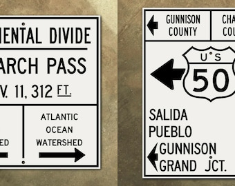 May include: A white sign with black text indicating the Continental Divide at Monarch Pass, elevation 11,312 feet. The sign also shows the Pacific and Atlantic Ocean watersheds. A second white sign with black text shows a US Route 50 sign with arrows pointing left and right. The sign also shows mile markers for Salida, Pueblo, Gunnison, and Grand Junction.