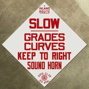 May include: A white diamond-shaped road sign with red text that reads "INLAND ROUTE SLOW GRADES CURVES KEEP TO RIGHT SOUND HORN" and a red circle with the text "AUTO CLUB SO. CAL."