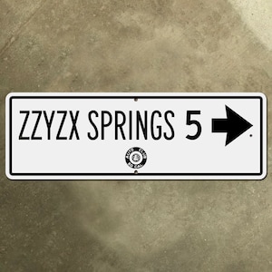 May include: White rectangular sign with black text and a black arrow pointing to the right. The sign reads "ZZYZX SPRINGS 5". A black and white circular sticker with the text "AUTO CLUB SO CAL" is in the lower right corner of the sign.