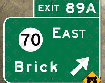 Könnte beinhalten: Grünes Straßenschild mit weißem Text und einem weißen Pfeil. Das Schild lautet "70 East Brick" und "Ausfahrt 89A". Ein gelbes Schild mit einem schwarzen Bären befindet sich in der unteren rechten Ecke.