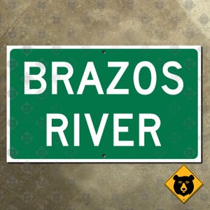 Könnte beinhalten: Ein rechteckiges grünes Schild mit den Worten "BRAZOS RIVER" in großen weißen Großbuchstaben. Das Schild hat einen Rand und ist auf einer strukturierten Oberfläche montiert. Ein gelbes rautenförmiges Schild mit einer schwarzen Bärgrafik befindet sich in der unteren rechten Ecke.