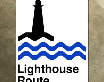 Op de afbeelding: Wit rechthoekig bord met een zwart silhouet van een vuurtoren boven blauwe golvende lijnen, met de woorden "Lighthouse Route" in het zwart. Het bord heeft een gat aan de bovenkant om op te hangen.