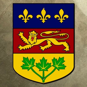 Puede incluir: Un emblema en forma de escudo con un esquema de colores azul, rojo y amarillo. La sección superior presenta tres flores de lis doradas sobre un fondo azul. La sección media representa un león dorado con una lengua roja sobre un fondo rojo. La sección inferior muestra tres hojas de arce verdes sobre un fondo amarillo.