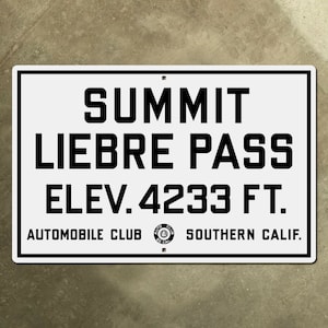 May include: White metal sign with black text that reads "SUMMIT LIEBRE PASS ELEV. 4233 FT. AUTOMOBILE CLUB SOUTHERN CALIF."