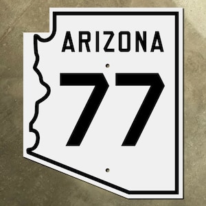 Puede incluir: Una señal de carretera blanca de Arizona con texto negro y borde. La señal muestra el nombre del estado "Arizona" y el número de ruta "77".