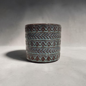 May include: A cylindrical ceramic pot with a textured surface. The pot features a repeating pattern of teal and brown chevrons. The pot is a decorative item, suitable for holding small plants or other items.