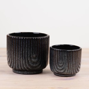 May include: Two black ceramic planters with a textured, ridged design. The larger planter has a wider base and a taller profile than the smaller one. Both have a dark, speckled finish and are suitable for indoor or outdoor use.