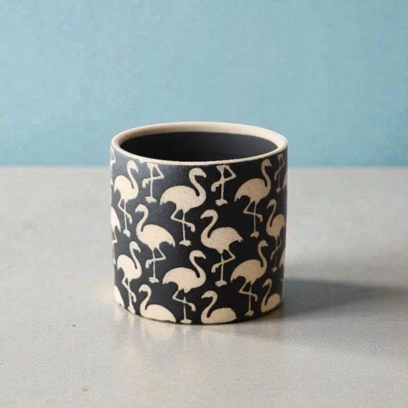 May include: A cylindrical ceramic planter with a black background and a repeating pattern of cream-colored flamingos. The interior is black, and the rim is cream-colored. The planter is suitable for small plants or decorative use.