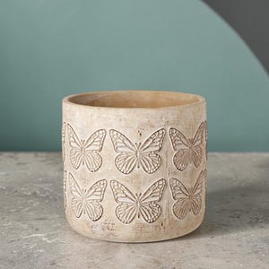 Op de afbeelding: Een beige cilindrische bloempot met een herhaald patroon van in reliëf gemaakte vlinders. De pot heeft een getextureerd oppervlak en is geschikt voor binnen of buiten. De vlinders zijn gedetailleerd met zichtbare vleugelpatronen.