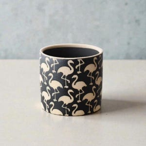 May include: A cylindrical ceramic planter with a black background and a repeating pattern of cream-colored flamingos. The interior is black, and the rim is cream-colored. The planter is suitable for small plants or as a decorative item.