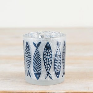 Puede incluir: Un portavelas de cristal esmerilado con interior plateado. El exterior presenta un patrón repetitivo de peces azules estilizados. Los diseños de los peces varían, con patrones que incluyen líneas onduladas, puntos y sombreado cruzado. El portavelas es cilíndrico.