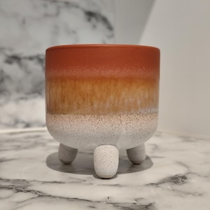 Pode incluir: Um pequeno vaso redondo de cerâmica com um gradiente de cores. A parte superior é vermelho-alaranjada, passando para castanho claro e depois branco na base. Possui três pernas curtas e arredondadas. Ideal para pequenas plantas ou suculentas.