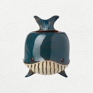 Op de afbeelding: Een blauwgroene keramische walvisvormige plantenbak met een glanzende afwerking. De plantenbak heeft een getextureerde, witte mond met verticale lijnen, kleine zwarte ogen en een gedetailleerde staartvin. Kleine vinnen bevinden zich aan de basis.