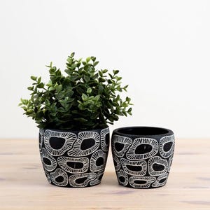 Pode incluir: Duas floreiras de cerâmica preta com um padrão abstrato branco. Uma floreira contém uma planta verde com folhas. As floreiras são cilíndricas e assentam sobre uma superfície de madeira clara. O desenho repete-se em ambas as floreiras.