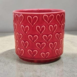 Peut inclure: Un pot de fleurs en céramique rouge avec un motif de cœurs répété. Le pot est cylindrique avec un bord légèrement évasé et une petite base. Les cœurs sont en relief, créant une surface texturée.