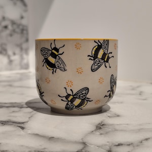 Könnte beinhalten: Eine cremefarbene Keramikschale, verziert mit schwarz-gelben Bienenillustrationen und kleinen orangefarbenen Blumenakzenten. Der Rand der Schale hat einen gelben Rand. Die Schale wird wahrscheinlich zum Servieren von Speisen oder als Dekorationsartikel verwendet.