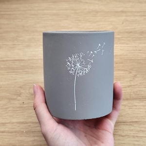 Op de afbeelding: Een grijze cilindervormige bloempot met een wit paardenbloemontwerp. De paardenbloem wordt afgebeeld met zijn zaden die in de wind waaien. De pot wordt in een hand gehouden en staat op een houten oppervlak.