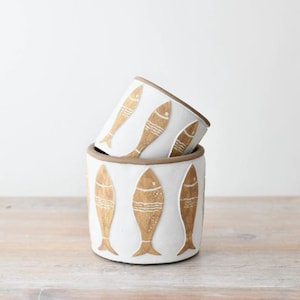 May include: Two white ceramic pots stacked, decorated with a repeating pattern of gold-coloured fish. The rims and bases of the pots have a brown trim. The pots are set on a light-coloured wooden surface.