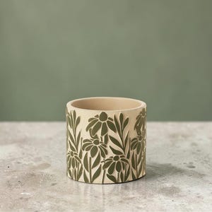May include: A beige ceramic planter with a floral design. The planter features a repeating pattern of green flowers and leaves around its exterior. The cylindrical pot is suitable for small plants and adds a touch of nature to any space.