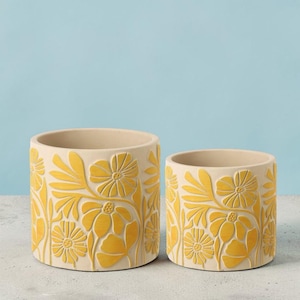 Op de afbeelding: Twee beige keramische bloempotten met een geel bloemenontwerp. De potten zijn cilindrisch, met de grotere aan de linkerkant en de kleinere aan de rechterkant. Het bloemenpatroon is in reliëf op het oppervlak.
