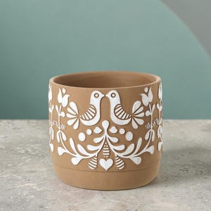 May include: A terracotta-coloured ceramic planter with a white embossed design featuring birds, flowers, and hearts. The planter has a cylindrical shape and sits on a textured surface. The design is symmetrical and detailed.