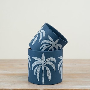May include: Two navy blue ceramic plant pots stacked on a wooden surface. Each pot features a white palm tree design around the exterior. The pots are cylindrical and have a smooth finish. The background is a neutral colour.