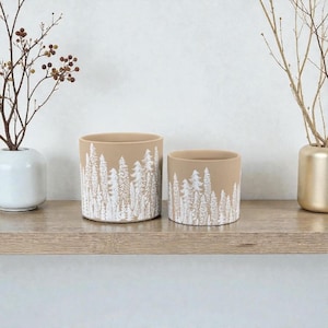 May include: Two beige ceramic plant pots with a white forest design. The pots are cylindrical, one larger than the other. They sit on a wooden shelf with decorative branches in vases on either side.