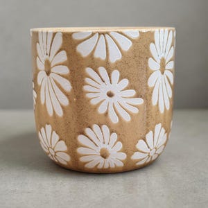 May include: A cylindrical, beige ceramic planter with a repeating pattern of white daisy flowers. The planter has a speckled texture and a smooth, rounded rim. The daisies are evenly spaced around the exterior.