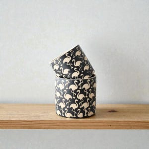 May include: Two cylindrical containers stacked on a wooden shelf. The containers are black with a repeating pattern of cream-colored flamingos. The containers are likely ceramic or similar material.