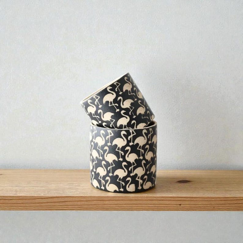 May include: Two cylindrical containers stacked on a wooden shelf. The containers are black with a repeating pattern of cream-colored flamingos. The containers are likely ceramic or similar material.