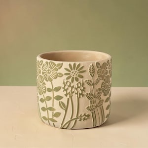 May include: A cylindrical, cream-coloured ceramic planter featuring a floral design in shades of green. The embossed flowers and foliage create a textured surface. Suitable for indoor or outdoor use.