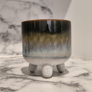 May include: A ceramic planter with a gradient glaze in shades of brown, green, and white. The planter has a cylindrical shape and sits on three small, rounded legs. The top rim is a dark brown color.