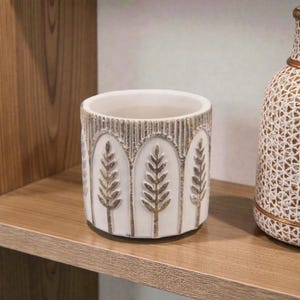 Könnte beinhalten: Weißer Keramik-Pflanztopf mit zylindrischer Form. Er hat ein sich wiederholendes Muster aus goldfarbenen Weizenähren auf weißem Hintergrund. Der Rand und die Oberkante haben ein strukturiertes, goldfarbenes Band. Geeignet für den Innenbereich.