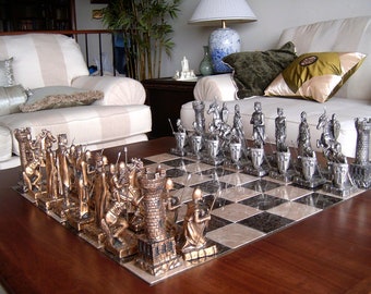 May include: A silver and bronze chess set with a marble chessboard. The chess pieces are in the style of medieval knights and are arranged on a wooden table.