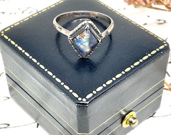 May include: A silver ring with a square, iridescent stone set in a diamond-shaped frame. The ring is displayed on a black jewelry box with gold trim and a gold clasp. The stone reflects blue, green, and gold hues.