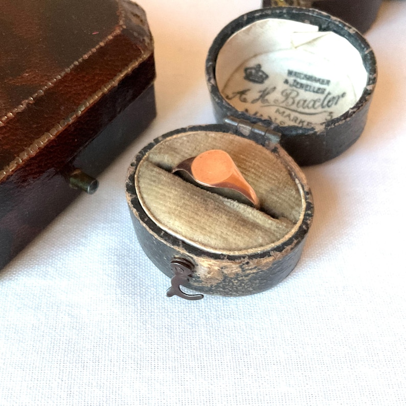 May include: A vintage signet ring with a peach-colored oval face, displayed in a small, antique leather ring box with a velvet interior. The box is open, revealing the ring. Other boxes are visible.
