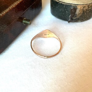 May include: A gold-colored ring with a triangular top and a circular band. The top of the ring has the letters "OCT" engraved on it. The ring is set against a white background, with a glimpse of a brown jewelry box.