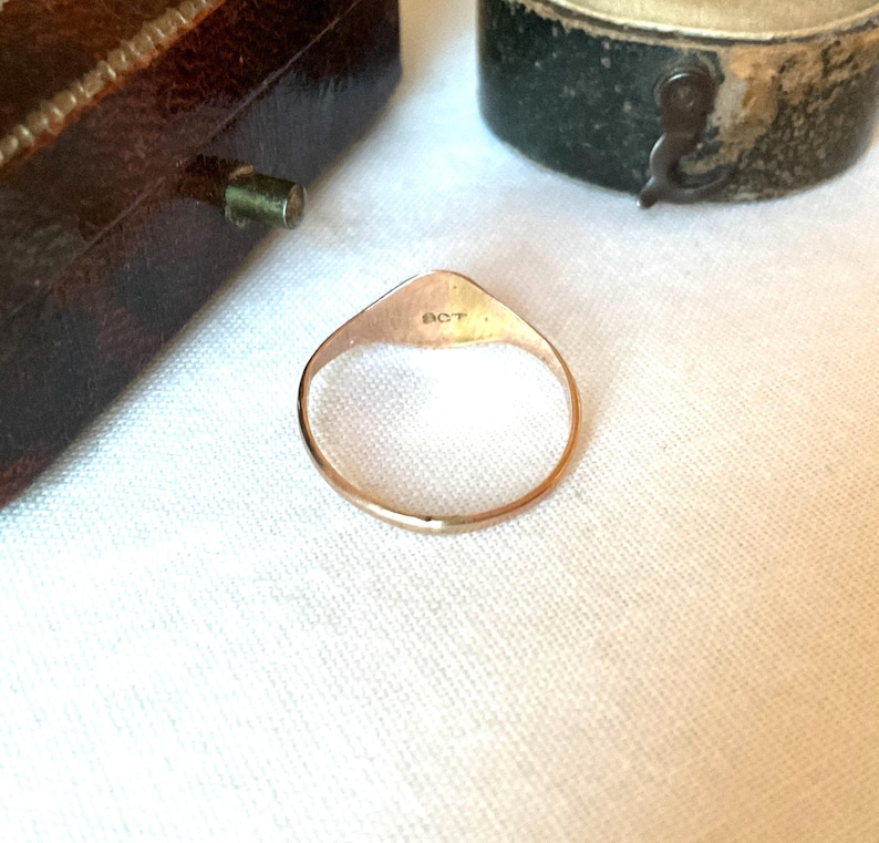 May include: A gold-colored ring with a triangular top and a circular band. The top of the ring has the letters "OCT" engraved on it. The ring is set against a white background, with a glimpse of a brown jewelry box.