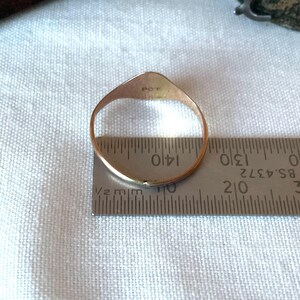 May include: A gold ring with a flat, shield-shaped top is measured with a ruler. The ring is placed on a white fabric. The ruler shows measurements in inches and millimeters.