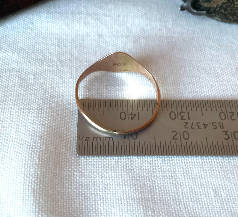 May include: A gold ring with a flat, shield-shaped top is measured with a ruler. The ring is placed on a white fabric. The ruler shows measurements in inches and millimeters.