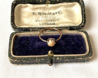 May include: A thin metal ring featuring a single round, pale pinkish-white pearl set in a four-prong mounting. The ring is displayed inside a rectangular, dark green, hinged jewelry box with a deep blue velvet interior and cream silk lining.
