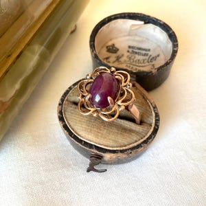 May include: A gold ring featuring a central oval cabochon-cut deep purple stone surrounded by a decorative openwork floral metal setting. The ring is displayed inside a round, dark-colored vintage jewelry box with a beige velvet interior lining.