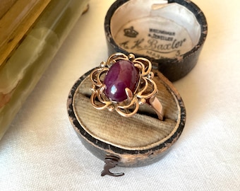 May include: A gold ring featuring a central oval cabochon-cut deep purple stone surrounded by a decorative openwork floral metal setting. The ring is displayed inside a round, dark-colored vintage jewelry box with a beige velvet interior lining.