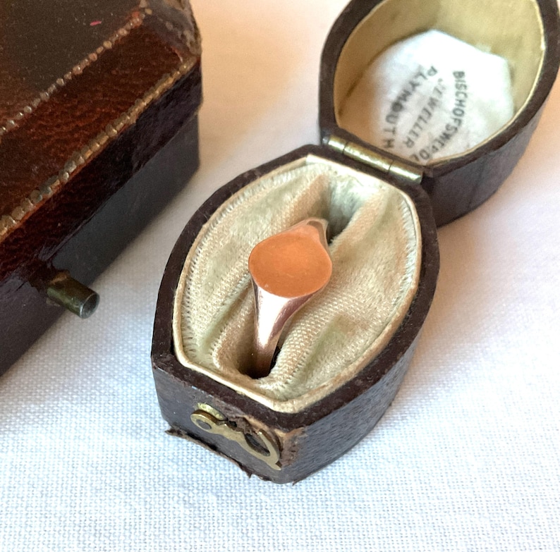 May include: A rose gold signet ring with a smooth, oval face, displayed in a vintage brown leather jewelry box with a cream-colored interior. The ring is set against a light beige fabric.
