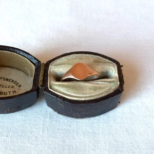 May include: A gold signet ring with a smooth, oval face rests inside an open, antique jewelry box. The box is dark brown with a light green interior. The text "BISCHOFSWERDER, JEWELLER, PLYMOUTH" is printed inside the box.