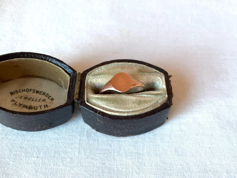 May include: A gold signet ring with a smooth, oval face rests inside an open, antique jewelry box. The box is dark brown with a light green interior. The text "BISCHOFSWERDER, JEWELLER, PLYMOUTH" is printed inside the box.