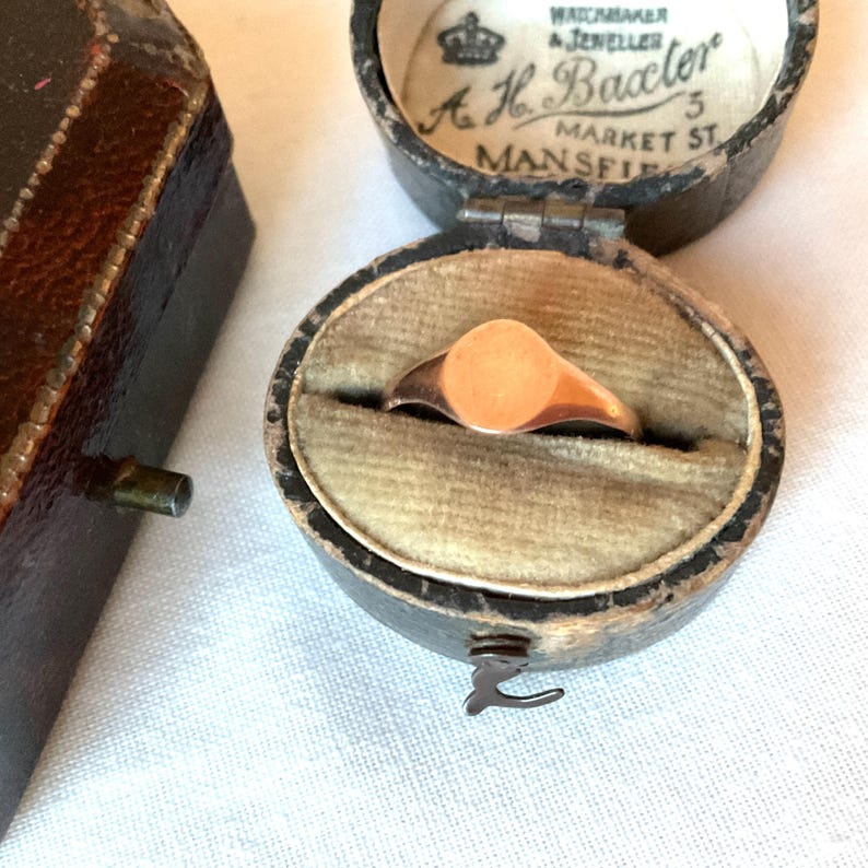 May include: A gold signet ring with a circular face rests inside a vintage black jewelry box with a velvet interior. The box is open, revealing the ring's design. The box has the text "Watchmaker & Jeweller A.H. Baaxter 5 Market St Mansfield".
